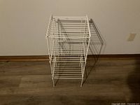 White steel wire rack with three shelves shown from front angle against wall on wooden floor.