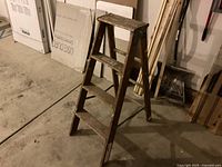 Wooden three step ladder standing unfolded on concrete floor, showing three steps per side and signs of wear on the wood.