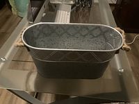 Front and side view of the oval tin basket showing metal with patterned sides and rope handles on a glass table.