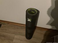 Standing vertical black TriggerPoint foam roller with ridges visible, against a wall on wooden floor.