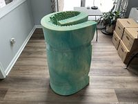 Photo showing rolled green foam mattress topper with textured surface for comfort and support, set on a hardwood floor with some boxes and furniture in the background.