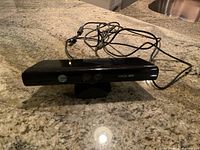 Black Xbox 360 Kinect sensor device placed on a granite surface, showing front with lenses and attached cable coiled behind.