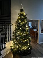 Photo of artificial Christmas tree set up indoors near staircase and hallway, lit with warm white string lights and decorated with silver baubles.