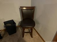 Front view of a single wooden barstool with a brown upholstered seat and backrest, placed against a beige wall and carpeted floor.