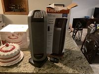 Front view of the Lasko ceramic tower heater along with its original box and remote control placed on a kitchen counter.