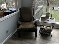 Photo of an upholstered armchair positioned near a window and side table with books and lamp. Fabric is patterned with muted brown tones; wooden arms and legs are dark and carved.