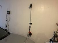 Photo showing the full weed wacker hanging on wall next to a car, highlighting the orange and black engine housing, long metal shaft, handle grip, and cutting guard.