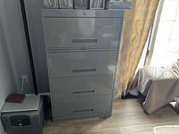 Photo showing the front view of a gray metal file cabinet with four drawers, lock on the top drawer, placed near a window with curtains and adjacent furniture.