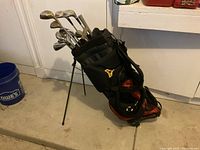 View of black and orange stand golf bag with golf clubs inside, showing extended legs and carrying straps.
