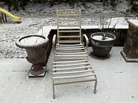 Single beige metal lounger and chaise lounge chair outdoors on a snowy surface, flanked by two planters and near a patio area.