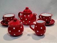 Wide view of entire 7-piece ceramic red and white polka dot tea set including teapot with lid, sugar bowl with lid, creamer, two cups, and two saucers.
