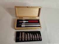 Top view of wooden box open showing three precision knives and one red handle knife, with black blade holder in front containing 12 assorted blades.