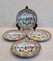Four large Chinese porcelain plates with colorful rooster designs, arranged with one on display stand