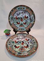 Two large Chinese porcelain plates, one standing and one lying facing up, showing rooster and floral designs with colorful borders.