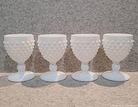 Full view of four hobnail milk glass goblets arranged in a row on a textured surface