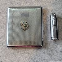 Photo showing the cigarette case with textured silver-tone finish and emblem on the front, alongside a slim silver-tone cigarette holder.