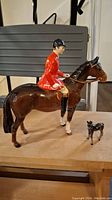 Mounted horse figurine with jockey in red jacket and small foal figurine on wooden surface.