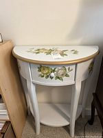 Full view of white demi lune table with floral decoration and single drawer, placed against wall indoors.