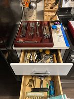 Wooden flatware organizer tray with forks, spoons, knives on countertop, open drawers below with more kitchen utensils visible