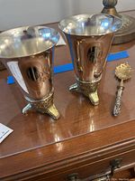 Photo showing two silver-tone cups with gold-tone tripod feet and one decorative spoon arranged on wooden surface, all tarnished and requiring cleaning.