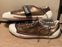 Side profile of the bronze and white Kappa sneakers with white soles and red stripe detail, showing upper materials and laces.