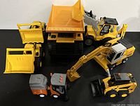 Seven toy construction vehicles arranged showing front and side details: includes dump truck, bulldozer, excavator, loader and street sweeper in yellow, black, orange and grey plastic.