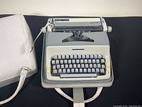 View of the 1960s Speedwriter Cavalier manual metal typewriter in gray and cream with portable white carrying case on black background.