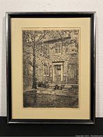 Framed black and white etching of a stone house facade under a tree, showing full artwork and frame.