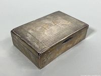 Top angled view showing rectangular shape, hinged lid, textured pattern, and monogram plaque with 'S'. Tarnish, dents, and wear visible.
