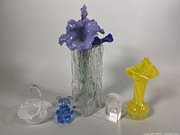 Photo showing full lot with textured vase, glass flower adornments, blue glass bear, clear glass basket, clear glass paperweight, and yellow glass vase.