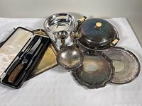 Photo of the vintage silver plated trays, covered serving dish, footed bowl, candleholders, and carving utensils arranged on a white cloth.