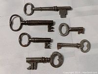 All six antique skeleton keys arranged on white fabric, showing different bow shapes and bit designs.