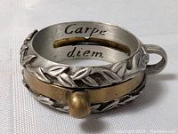Close-up image of a vintage metal pendant ring with 'Carpe diem' engraved inside and decorative leaf motif on the outside.