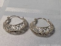 Pair of sterling silver hoop earrings laid on fabric surface showing intricate cut-out design and clasp closure.