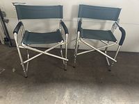 Front view of two lightweight aluminum camp chairs with green canvas seats and backrests, black padded armrests.