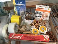 Photo showing various hardware items including rolls of masking tape, duct tape, tracing paper box, rope caulk box, wooden clothespins, coiled white cord, safety glasses, and small hardware pieces in a box.