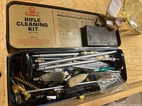 Open metal case showing contents of Outers Rifle Cleaning Kit including cleaning rods, brass and synthetic brushes, and patches.