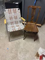 Two chairs on concrete floor: lawn chair with metal frame and woven patterned fabric, wooden chair with carved backrest