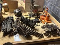 Photo showing grouped curved and straight black train tracks, transformer, water tower, telephone pole, rocket launching toy, and other accessories on a wooden surface.