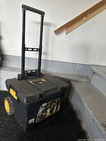 Black plastic rolling tool box with yellow wheels and extending handle, viewed from an angle showing the handle fully extended.