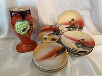 Photo shows orange and black hand painted Sakuraware vase, sugar bowl with landscape design, and stack of four saucers and five Nippon plates with landscape/pine tree scenery in orange gradient colors.