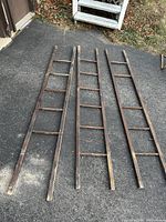 Three primitive wooden ladders laid flat on asphalt surface, showing entire length and rustic wear.