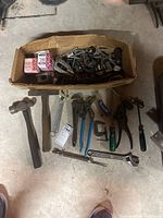 Box containing various hardware such as metal clamps, hinges, fasteners, and parts. Tools laid out in front include hammers, adjustable wrenches, pipe wrenches, pliers, two C-clamps, two screwdrivers, a ratcheting wrench, and a ratchet handle.