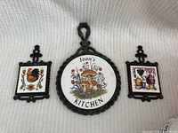 Three vintage kitchen trivets laid out on a white cloth background. Center is a round trivet with mushrooms and 'Joan's Kitchen' text, flanked by two smaller rectangle trivets with rooster and kitchen-themed ceramic tile images.