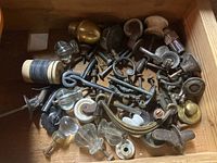 Top view of a wooden drawer filled with assorted vintage hardware pieces including clear glass knobs, brass knobs, and metal parts.