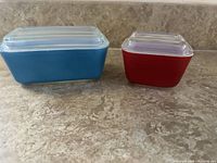 Two vintage Pyrex refrigerator dishes side by side on counter, one blue rectangular with lid and one red square with lid.