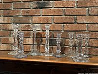 Eight glass or crystal candlesticks grouped together on a wooden surface in front of a brick wall, showing varied shapes and heights up to 10 inches.