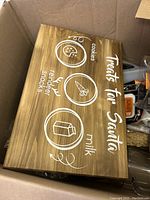 Wooden wooden sign with white icons and text "Treats for Santa" showing cookies, reindeer snacks, and milk