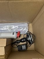 Box open showing black electronic device with attached power adapter and cables, rolled white sheets or posters, and screen cleaner can