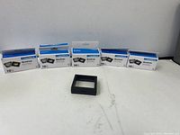 Six boxes of Leuchtturm Quadrum coin capsules with one capsule displayed in front on table. Capsules are square shaped with black frame.